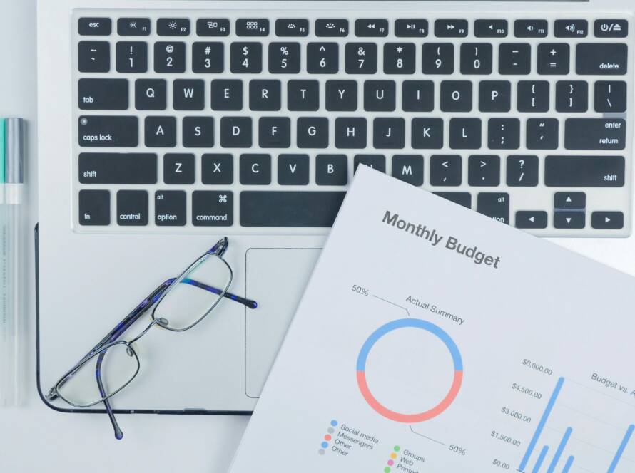 Top-down view of a laptop keyboard with a budget report and charts, glasses, and pastel markers on a white desk.