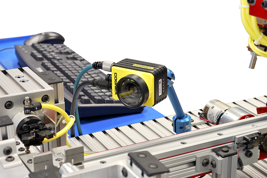 Industrial machine-vision camera (yellow Cognex) mounted over a conveyor belt with cables and robotics pieces nearby.