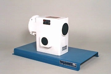 White cube-shaped industrial sensor mounted on a blue base plate, with two round black ports on the front and a small display on top.