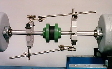 Lab shaft balance rig: a long metal shaft with green counterweights between adjustable silver clamps, mounted on a blue base with gray end disks.