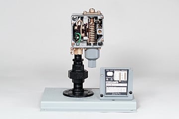 Desktop mechanical testing rig with exposed gears and wiring, mounted on a light gray base; a control panel sits to the right, illustrating a precision material tester.