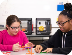Two girls wearing glasses build colorful 3D-printed models at a classroom table, with 3D printers in the background in a makerspace setup.