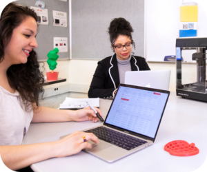 Two women collaborate at a desk with laptops; one writes on paper while a 3D printer sits nearby.