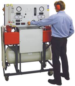Technician wearing earmuffs adjusts controls on a large industrial compressor test bench with gauges and meters.